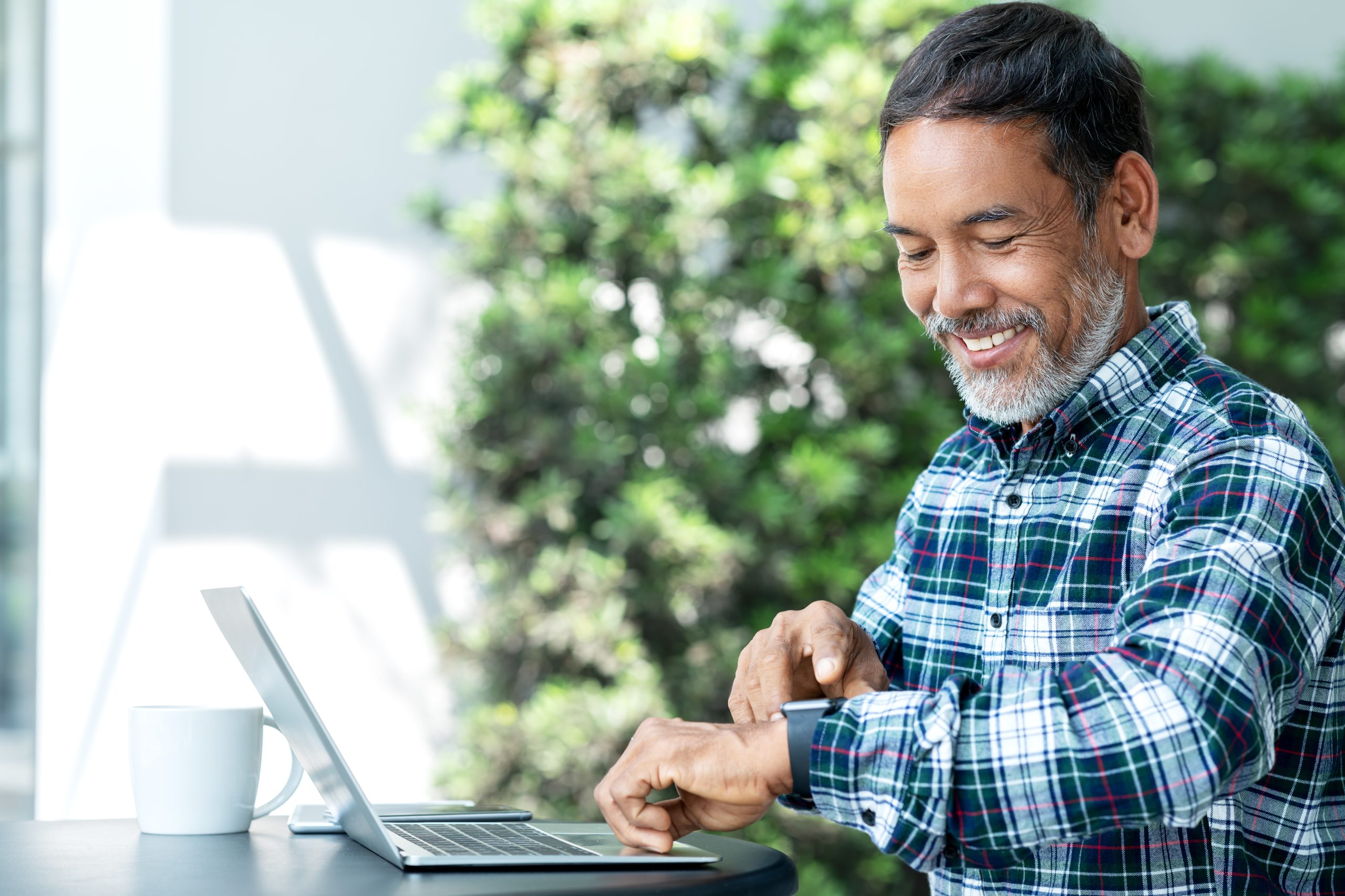 Smiling happy mature asian man with white stylish short beard using digital smartwatch and touching screen at coffee shop outdoor. Old indian or hispanic man using wearable technology with confident.